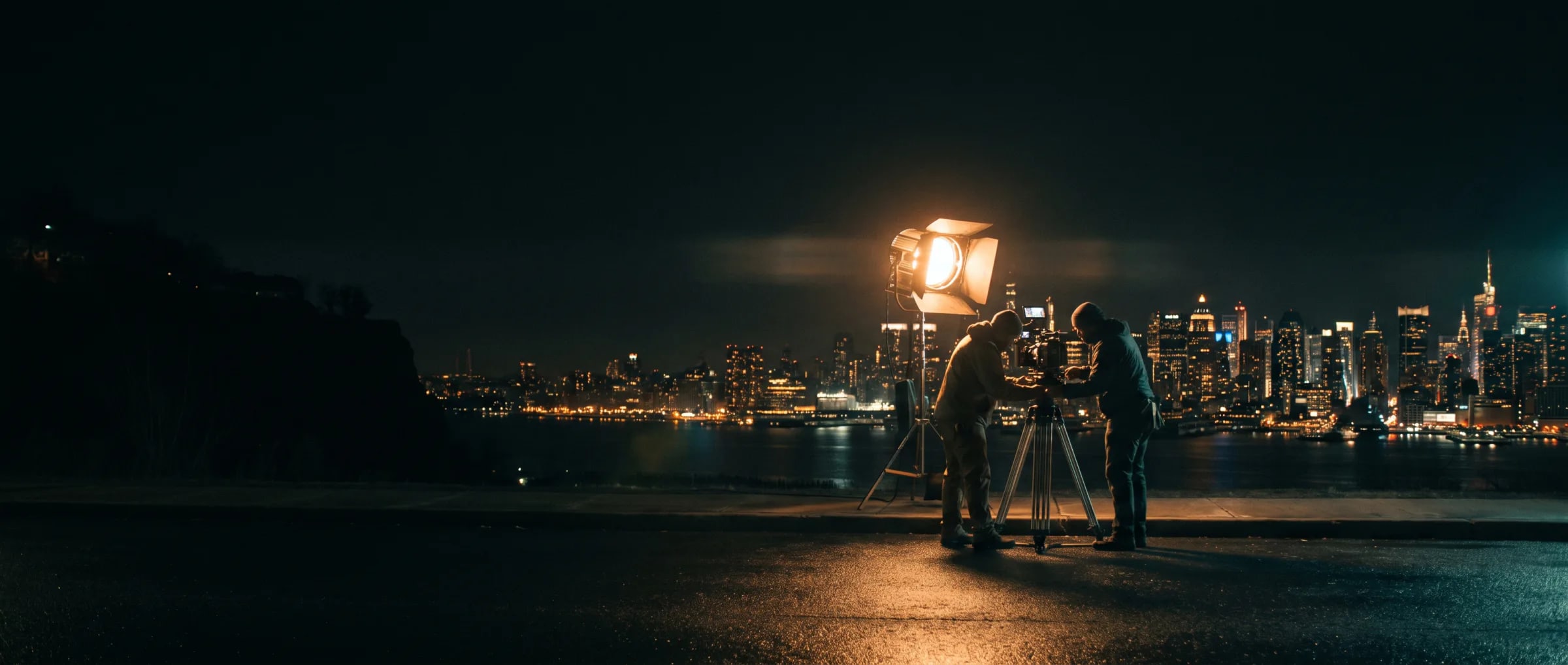 Night film shoot in New Jersey overlooking the Manhattan skyline across the Hudson — crew at a camera beside a lit fresnel